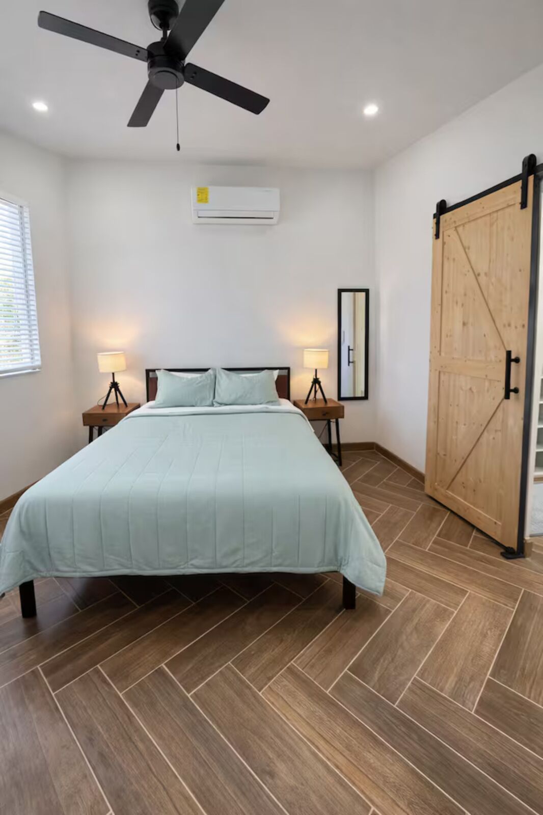 Une chambre à coucher comprenant un lit, deux lampes de chevet, un ventilateur de plafond, un appareil de climatisation, une porte en bois et un parquet à motifs en chevrons.