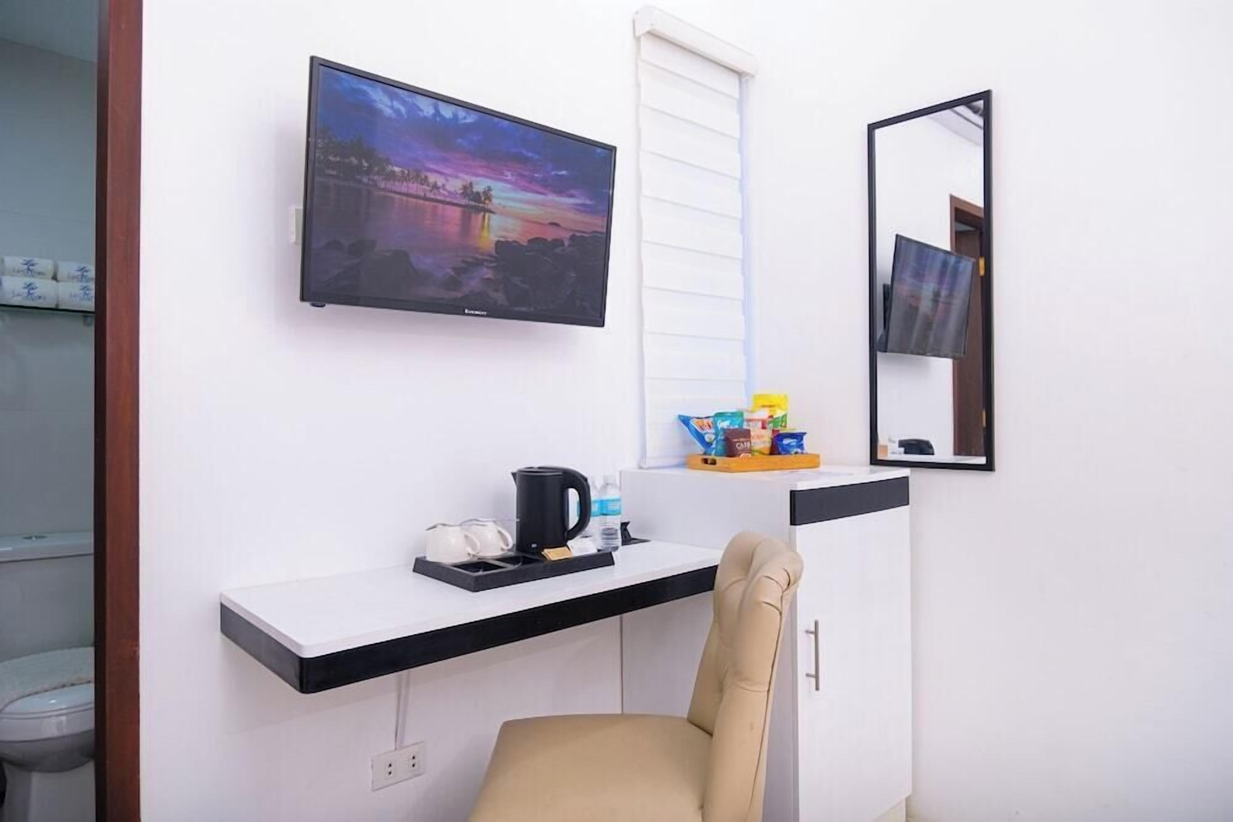 A room with a flat-screen TV mounted on the wall, a small coffee station, a chair, and a mirror.