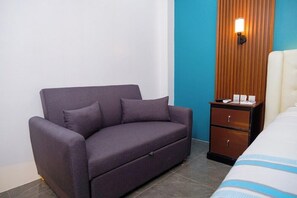 A room with a grey sofa, a wooden nightstand with a lamp, and a bed with a striped bedspread. - Le Dream Hotel (Vigan)