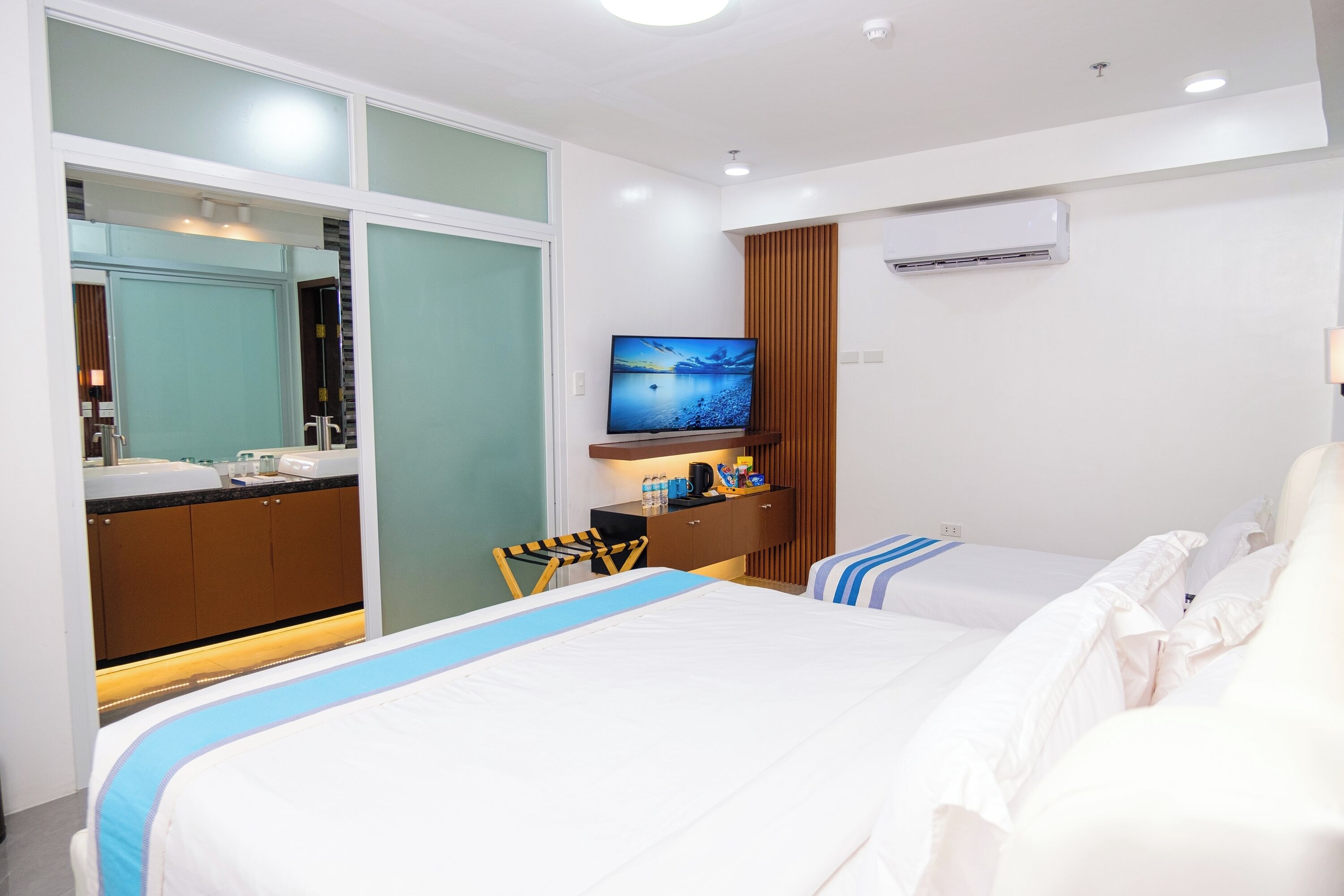 A hotel room with two beds, a TV, a desk with a mirror, and an air conditioning unit.