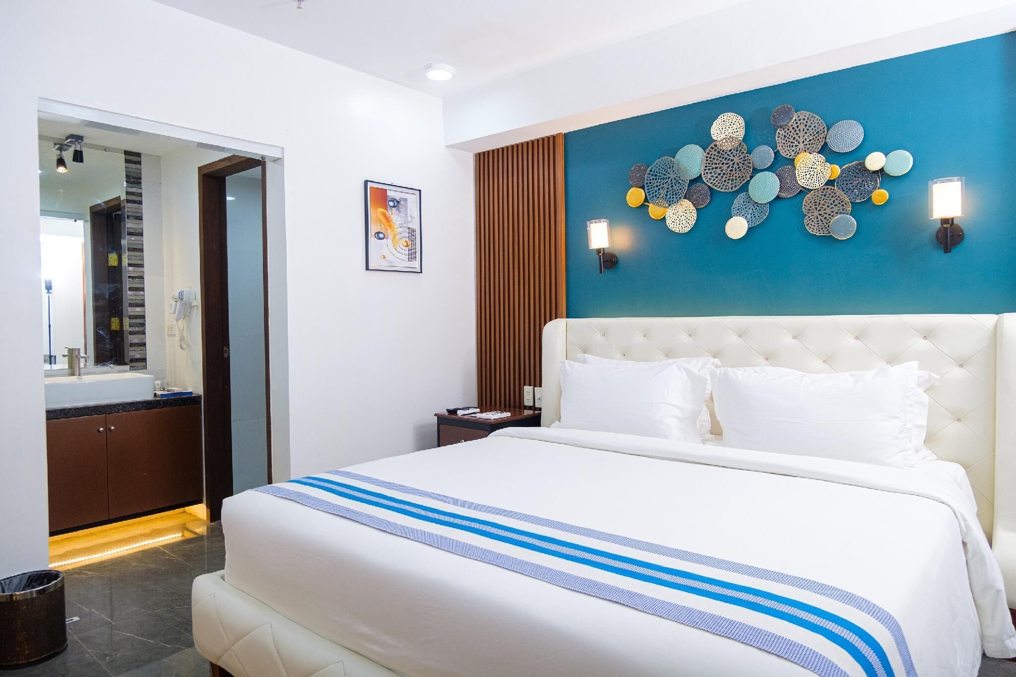 A hotel room with a bed, a nightstand, a wall-mounted lamp, a mirror, and a decorative wall piece.