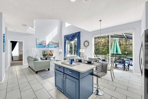 Eine moderne Küche mit Kochinsel, ein Essbereich mit Gartenmöbeln und ein Wohnzimmer mit Sofa und Fernseher.