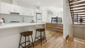 A modern kitchen with white cabinets, a central island with stools, and a staircase leading to a dining area. - Domi Rentals The Chadstone Homes (Chadstone)