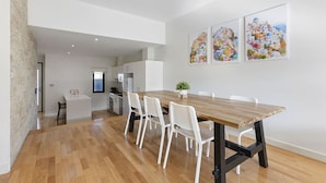 A spacious dining area with a wooden table and white chairs, a kitchen in the background, and three colorful paintings on the wall. - Domi Rentals The Chadstone Homes (Chadstone)