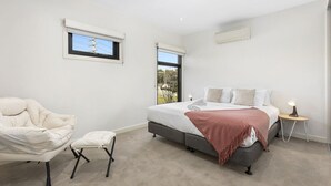 A bedroom with a bed, a chair, a small table, and a window. - Domi Rentals The Chadstone Homes (Chadstone)