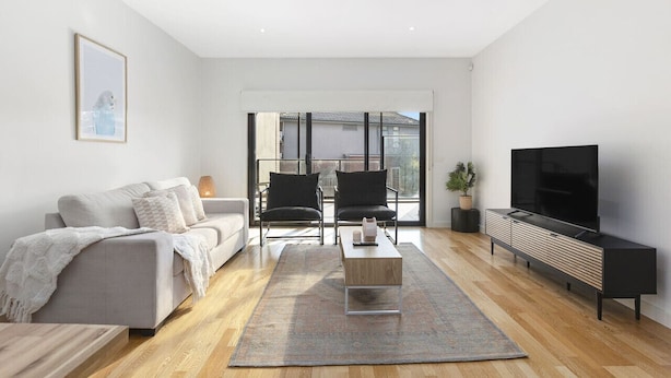 A modern living room with a sofa, chairs, a coffee table, a TV, and a large window. - Domi Rentals The Chadstone Homes (Chadstone)