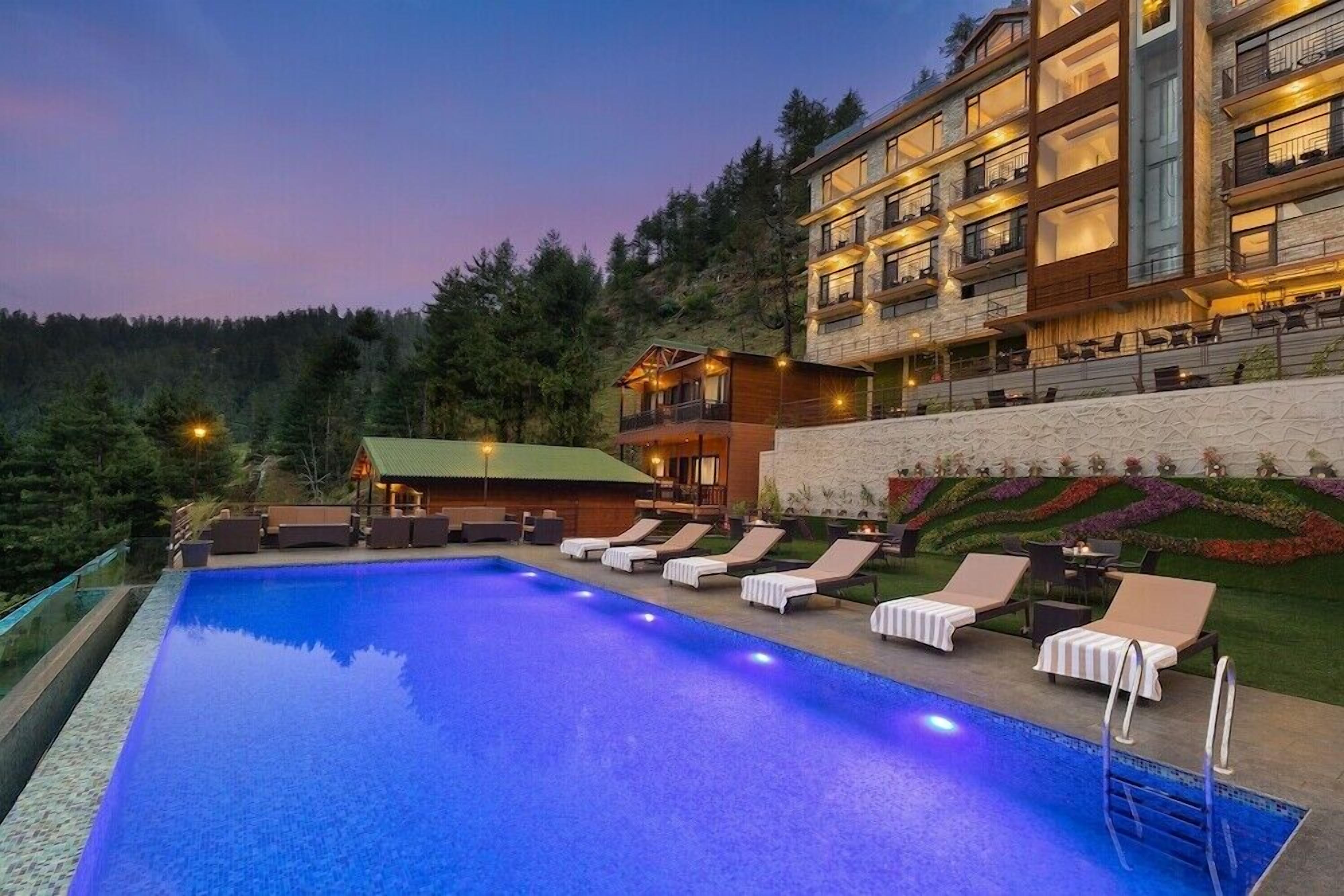 Una piscina con sillas de descanso, un edificio con balcones y un paisaje montañoso cubierto de bosque al fondo.