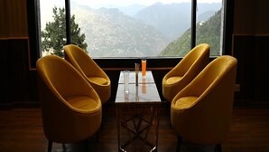 A room with two yellow chairs and a small table, a glass of orange drink, and a view of mountains through large windows. - Justa Luxe Cliffend Resort and Spa Mashobra (Shimla)