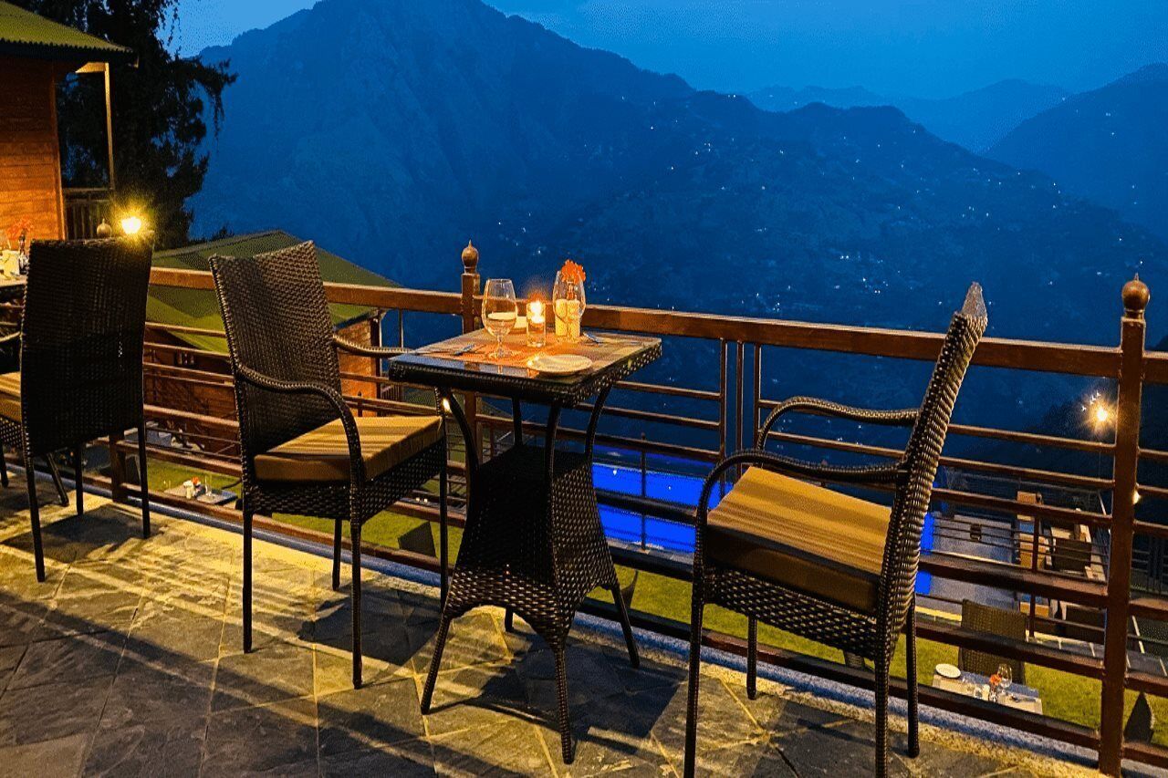 An outdoor dining area with wicker chairs and a table set for two, overlooking a mountainous landscape.