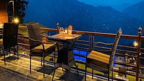 An outdoor dining area with wicker chairs and a table set for two, overlooking a mountainous landscape. - Justa Luxe Cliffend Resort and Spa Mashobra (Shimla)