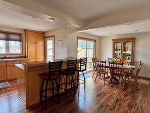 A spacious kitchen with wooden cabinets, a central island with bar stools, a dining area with wooden chairs, and large windows allowing natural light. - Lakefront Hot Tub Retreat w/ Mega Arcade (Baxter)