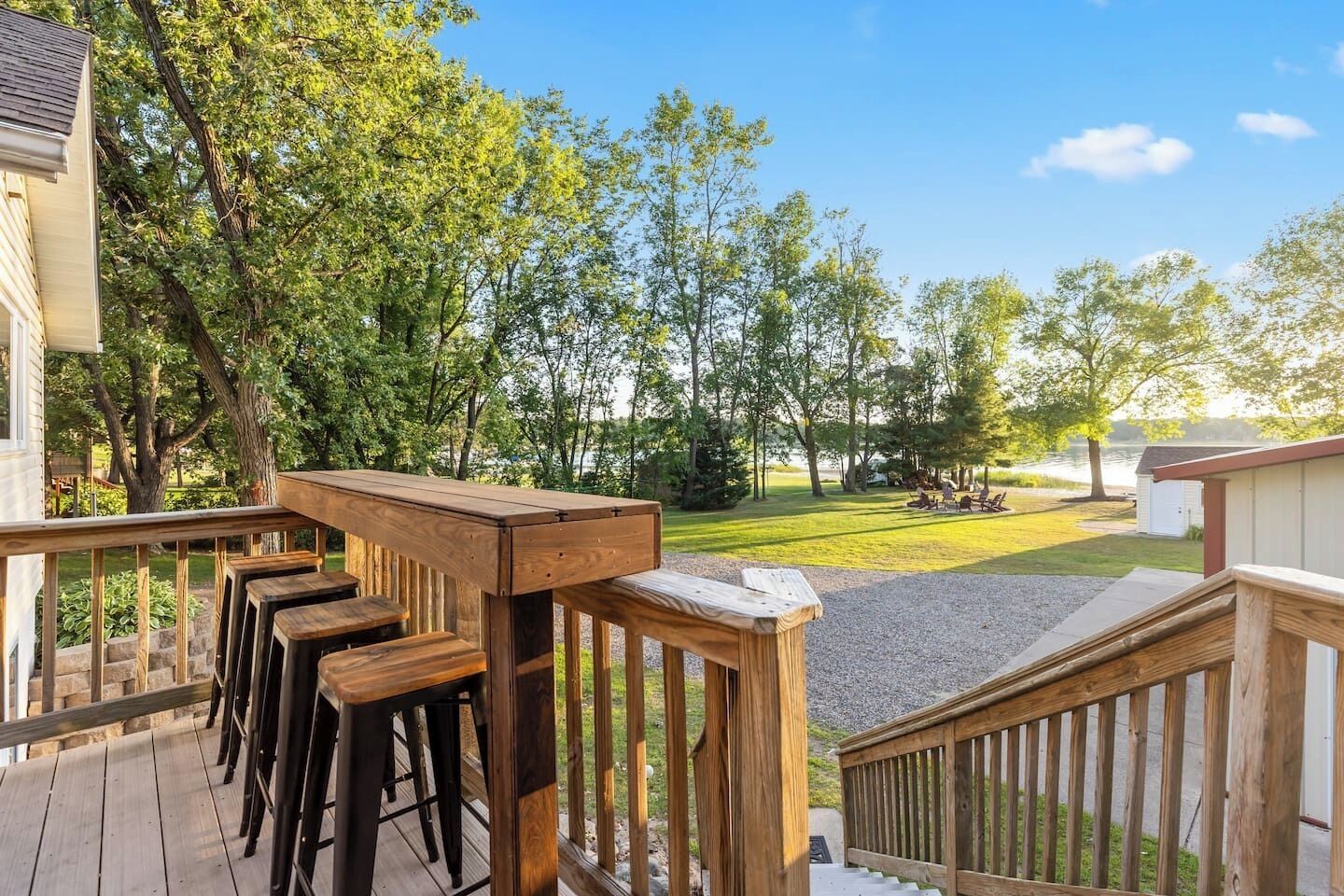 A wooden deck with a railing, a series of wooden stools, and a view of a grassy area with trees and a body of water in the background.