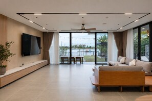 A modern living room with a flat-screen TV, a sofa, a dining table, and a view of a body of water. - GiaLinh Villa Hoi An - 5BR - River view (Hoi An)