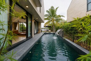 A swimming pool with a waterfall feature, surrounded by a modern building with large windows and outdoor seating. - GiaLinh Villa Hoi An - 5BR - River view (Hoi An)