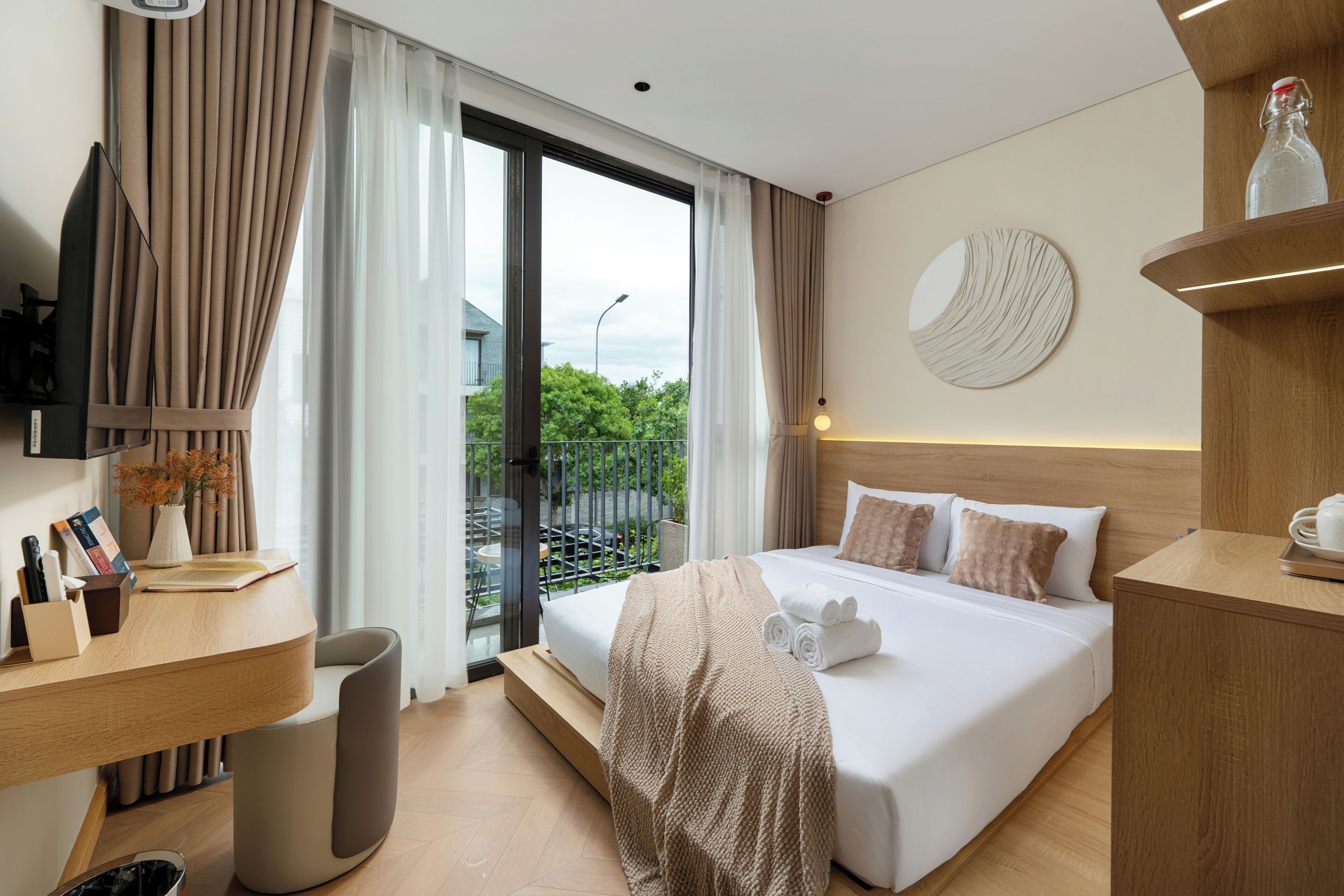 Quarto de hotel moderno com uma cama grande, uma escrivaninha com computador, vista para o exterior e um objeto decorativo redondo na parede.