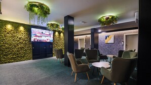 En lobby med et stort fladskærms-tv, siddeområde med stole og borde, dekorative planter og en væg med struktur.