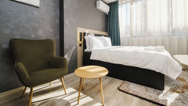 Une chambre d’hôtel moderne avec un lit simple, un fauteuil vert, un petit tabouret en bois et une grande fenêtre ornée de rideaux légers.