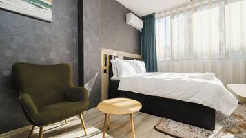 A modern hotel room with a single bed, a green armchair, a small wooden stool, and a large window with sheer curtains.