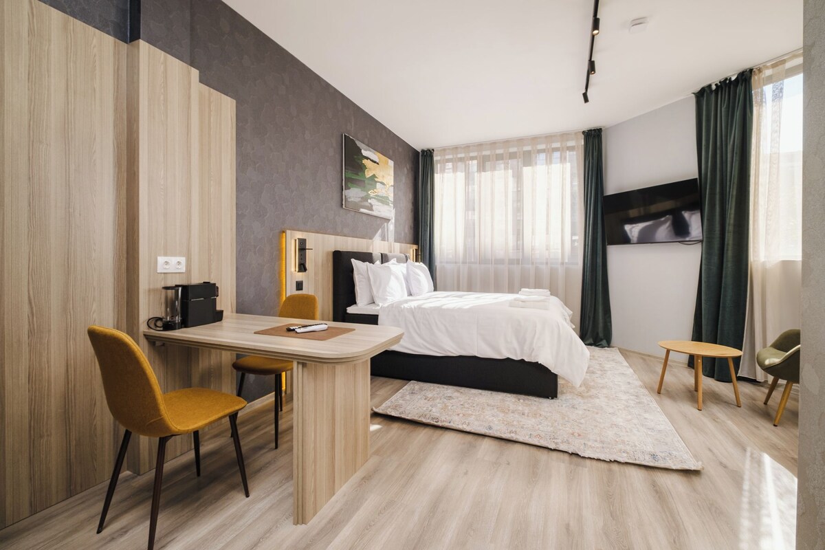 A modern hotel room with a bed, a desk, two chairs, a coffee maker, and a large window with curtains.
