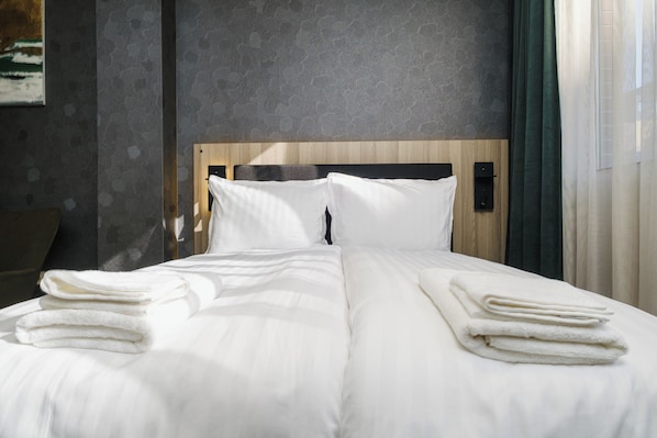Una cama bien tendida con sábanas blancas y toallas dobladas, un cabecero de madera y una pared texturizada de fondo.