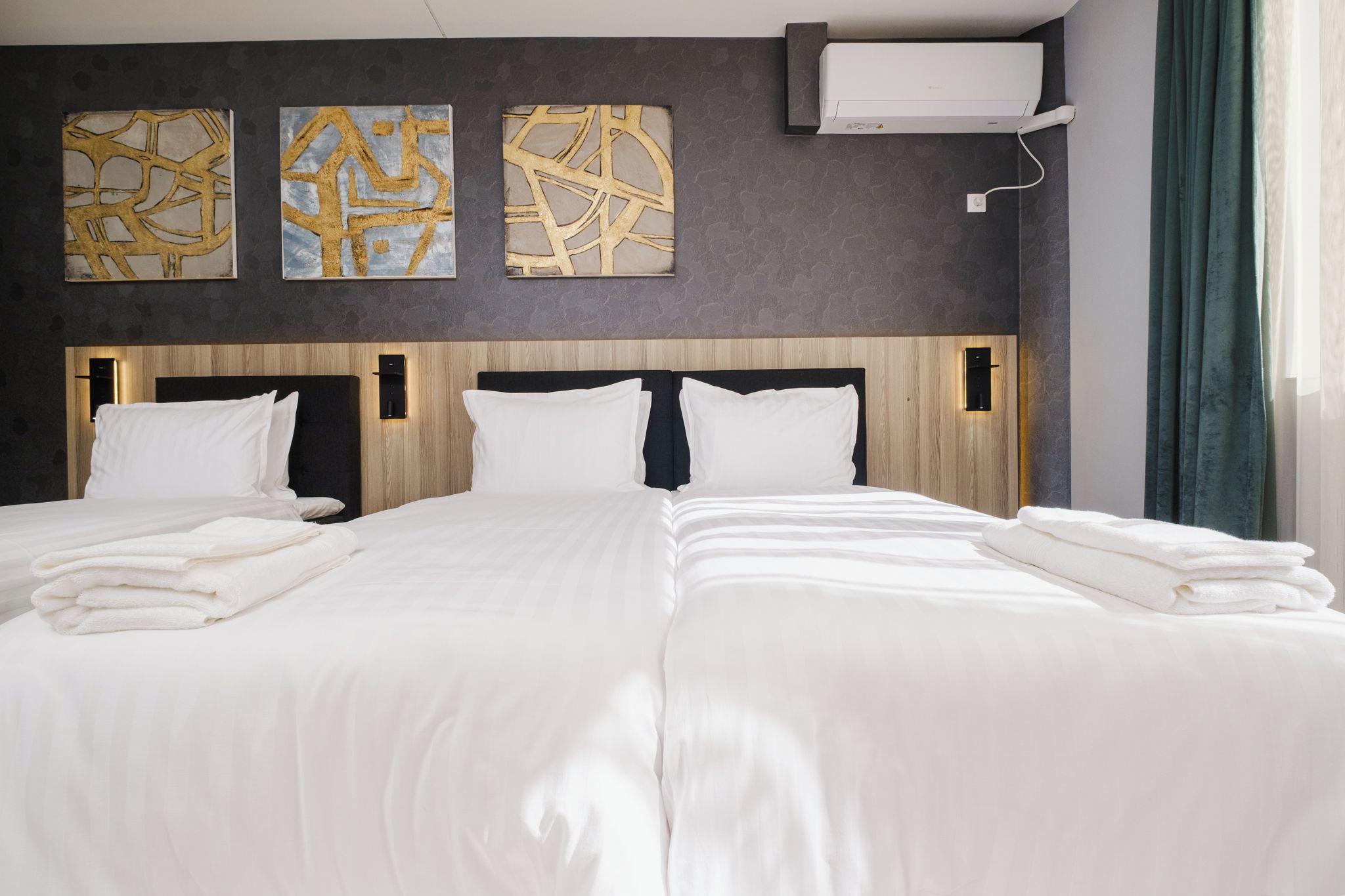 A double bed with white linens and towels, a wooden headboard, and three abstract gold and grey wall art pieces.