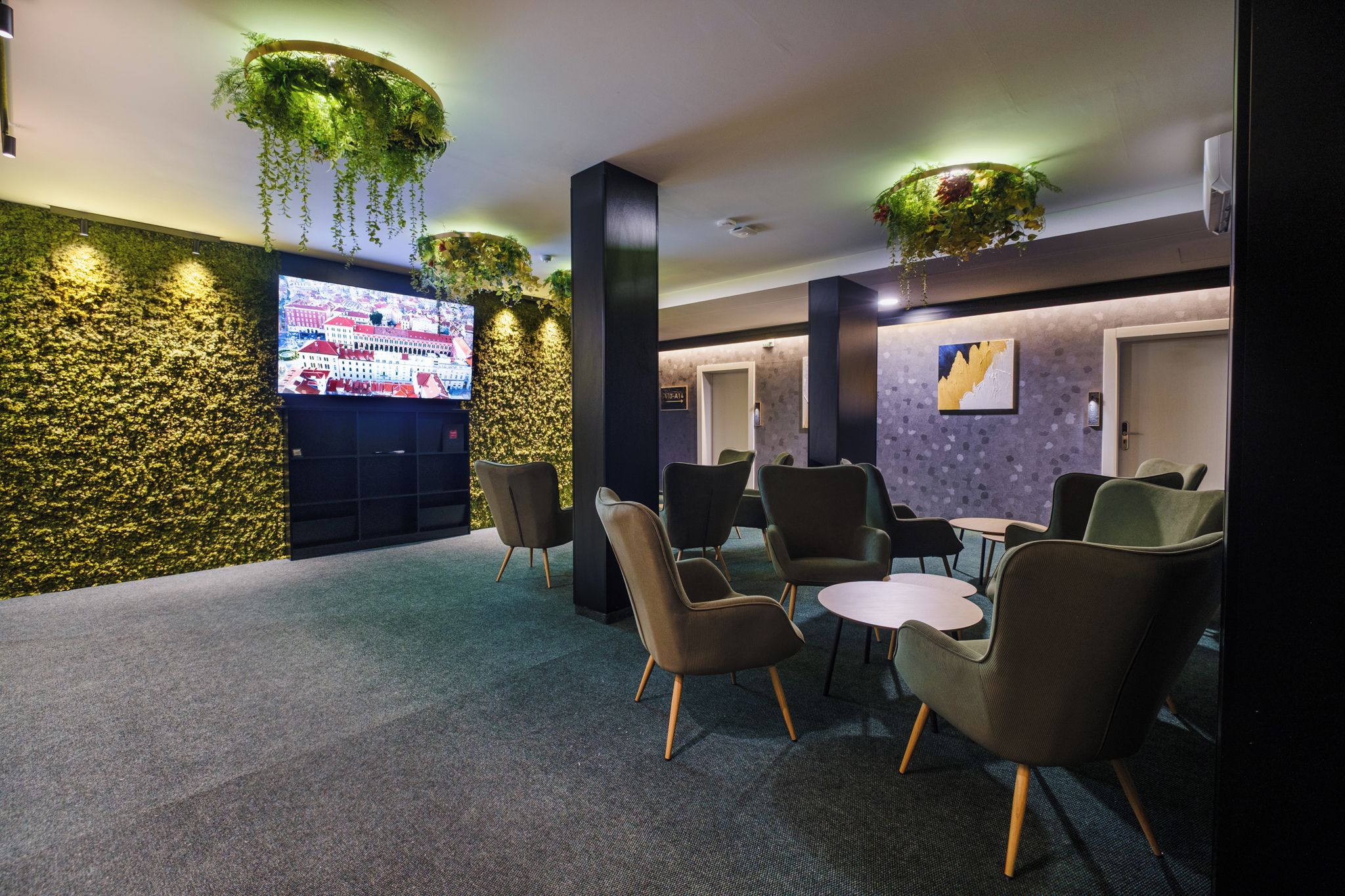 A lobby with a large flat-screen TV, seating area with chairs and tables, decorative plants, and a textured wall.