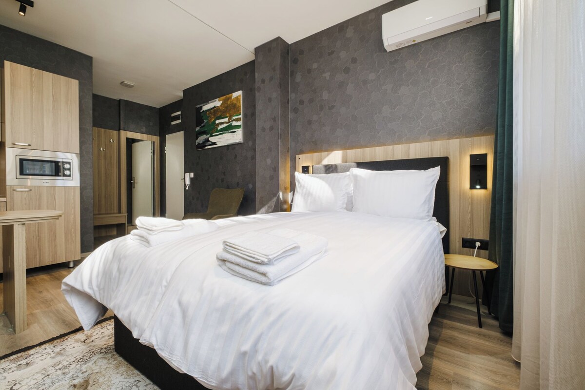 A modern hotel room with a large bed, a desk with a microwave, and a painting on the wall.
