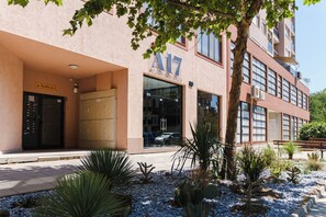 Un edificio con un letrero que dice 'A17' y 'Plovdiv Urban Suites'. Hay un estacionamiento con coches y un pequeño jardín con cactus y otras plantas desérticas.