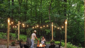 Un espacio al aire libre con sillas y un hogar de fuego, rodeado de árboles y luces colgantes.
