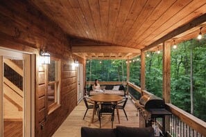 Eine Holzterrasse mit rundem Tisch und Stühlen, einem Grill und einer Couch mit Kissen.