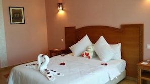 A hotel room with a neatly made bed, a teddy bear, and rose petals arranged on the bed.