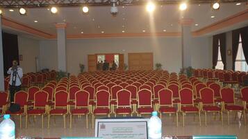 Un salón de conferencias con filas de sillas rojas, un escenario con una cámara y un atril con una placa.