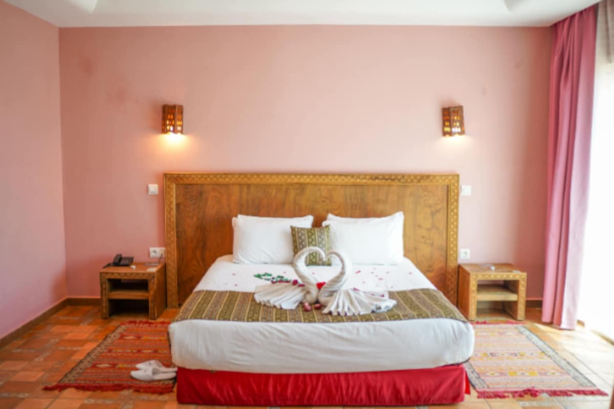 A hotel room with a large bed, two bedside tables, a wooden headboard, and a red skirt on the bed.