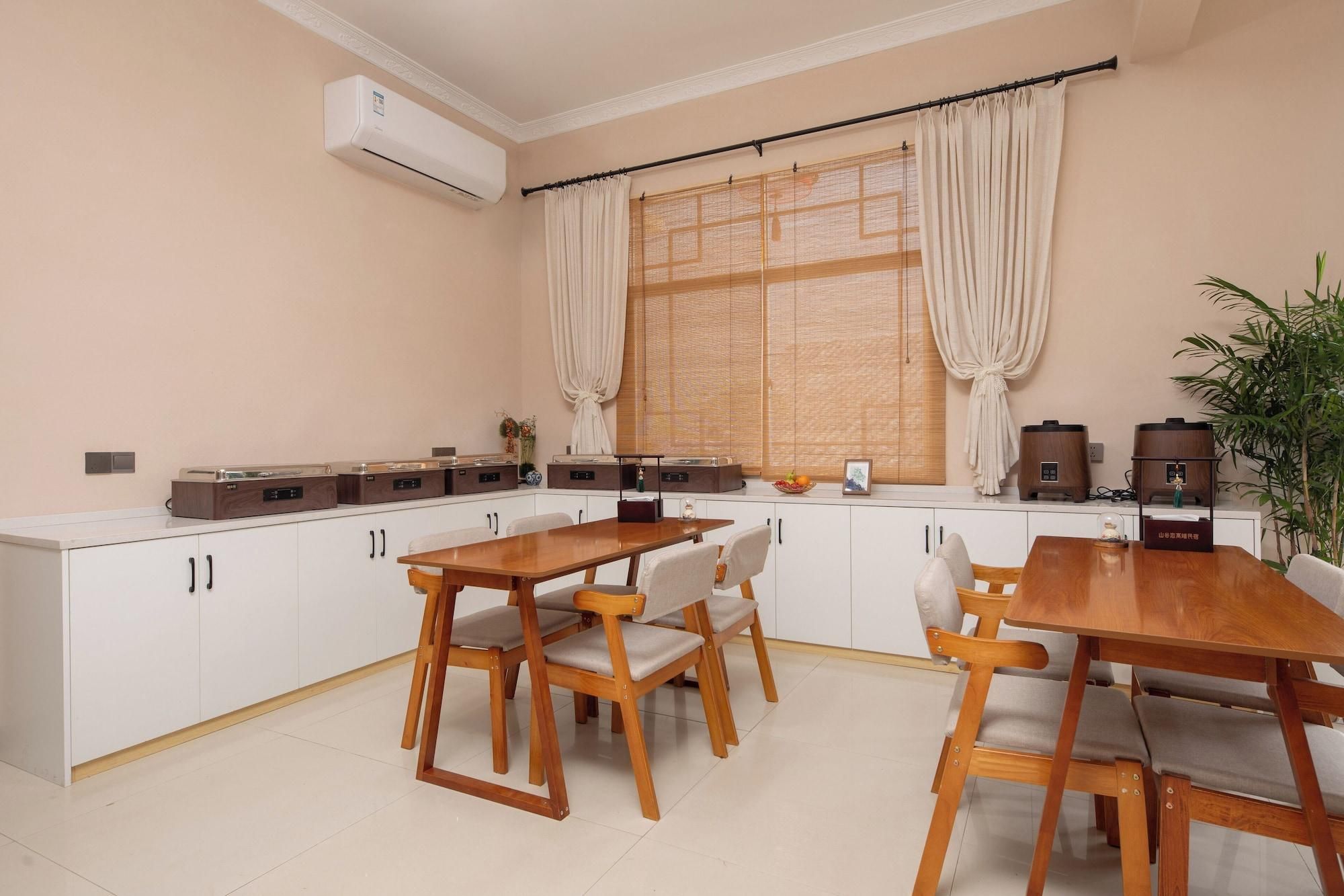 Een keuken met een eethoek, witte kasten, houten stoelen, een tafel, een airconditioner, een raam met gordijnen en een plant.