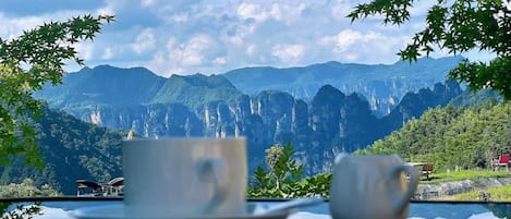 Una mesa con dos tazas y platillos blancos, con montañas y un cielo despejado de fondo.