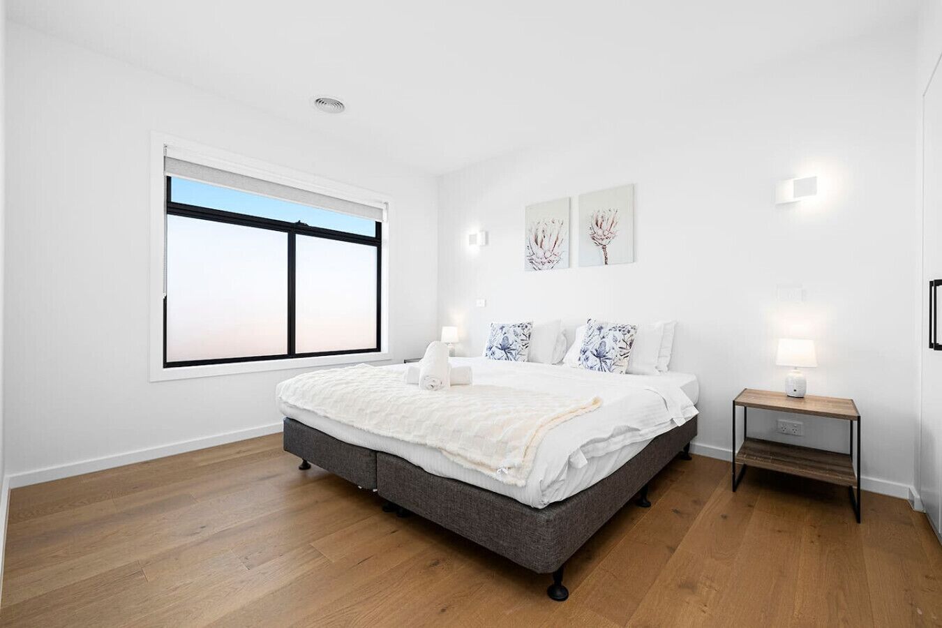 Une chambre avec un grand lit, une table de chevet avec une lampe, une fenêtre avec des stores et des œuvres d’art accrochées au mur.