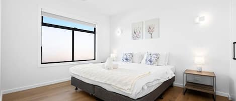 A bedroom with a large bed, a bedside table with a lamp, a window with blinds, and artwork on the wall.