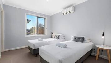 A hotel room with two beds, a window with a city view, an air conditioning unit, and a bedside table with a lamp.