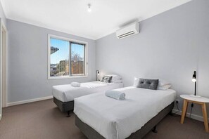A hotel room with two beds, a window with a city view, an air conditioning unit, and a bedside table with a lamp. - Domi Rentals - The Iona Townhouse (Clayton)