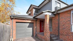 A two-story house with a garage, a red door, and a brick exterior. - Domi Rentals - The Lona Townhouse (Clayton)