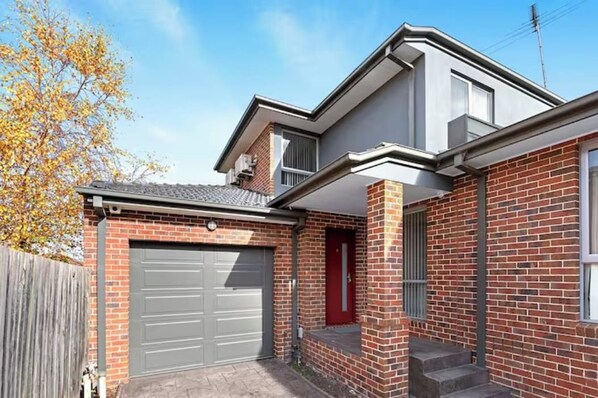 A two-story house with a garage, a red door, and a brick exterior. - Domi Rentals - The Iona Townhouse (Clayton)