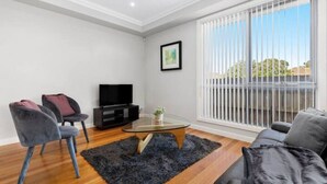 A modern living room with a flat-screen TV, two grey chairs, a glass coffee table, and a large window with blinds. - Domi Rentals - The Lona Townhouse (Clayton)
