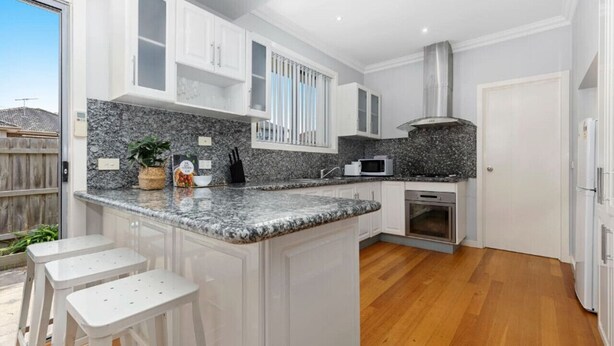 A modern kitchen with white cabinetry, granite countertops, and a central island with bar stools. - Domi Rentals - The Lona Townhouse (Clayton)