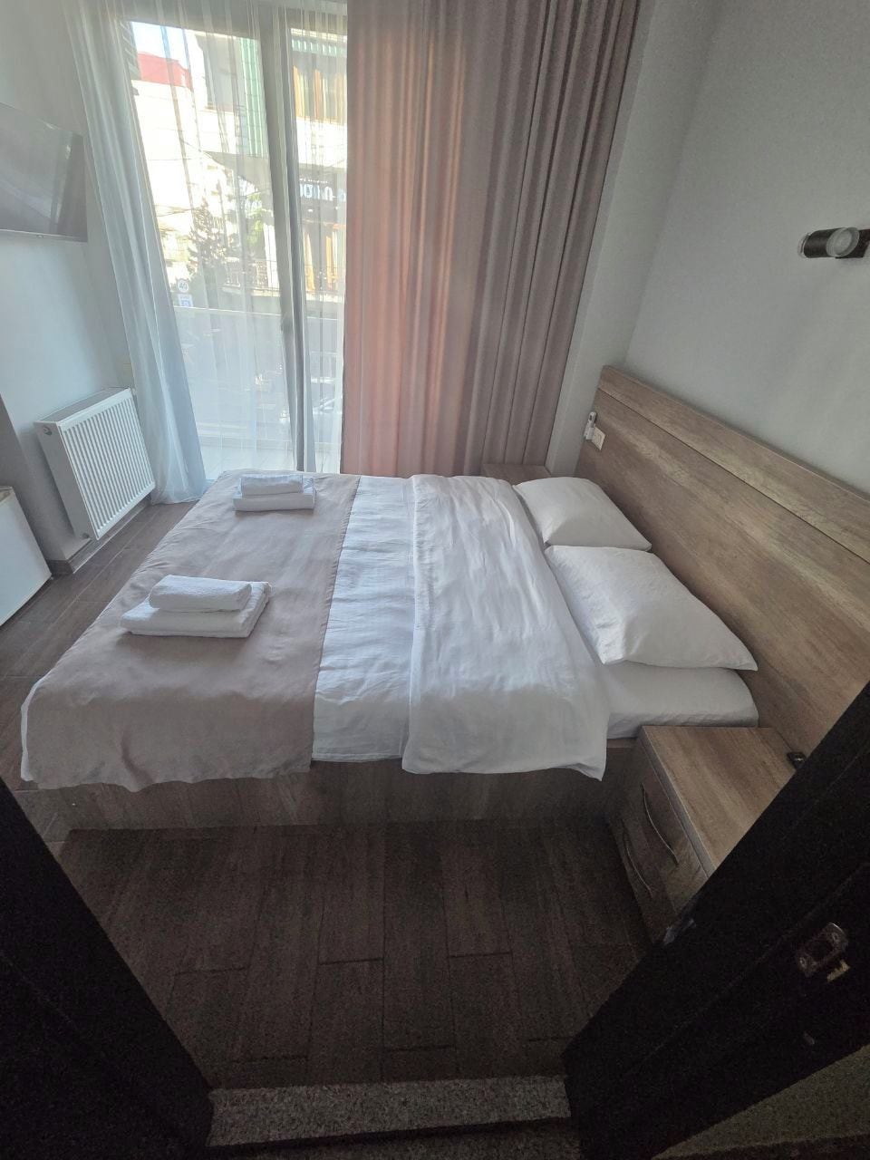 A hotel room with a bed, a wooden headboard, a window with curtains, and a radiator.
