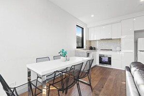 A modern kitchen with white cabinetry, a built-in oven, and a dining area with a white table and black chairs. - Domi Rentals - The Balaclava Apartment (Caulfield North)