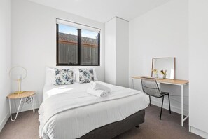 A bedroom with a bed, a desk, a chair, a window, and a small table with a lamp. - Domi Rentals - The Balaclava Apartment (Caulfield North)