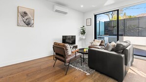 A modern living room with a sofa, armchair, TV, and artwork on the wall. - Domi Rentals - The Balaclava Apartment (Caulfield North)