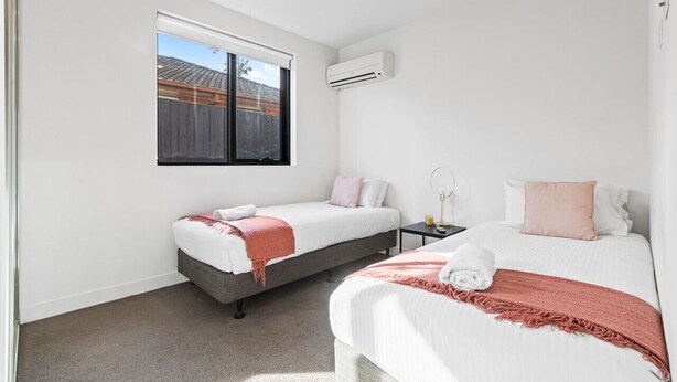 A bedroom with two beds, a window, an air conditioning unit, and a small bedside table with a lamp. - Domi Rentals - The Balaclava Apartment (Caulfield North)