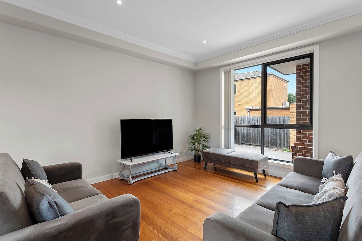 A modern living room with a flat-screen TV, grey sofas, a wooden floor, and a large window with a view of a brick building.
