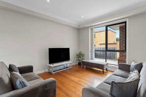 A modern living room with a flat-screen TV, grey sofas, a wooden floor, and a large window with a view of a brick building. - Domi Rentals - The Lantana Residence (Clayton)